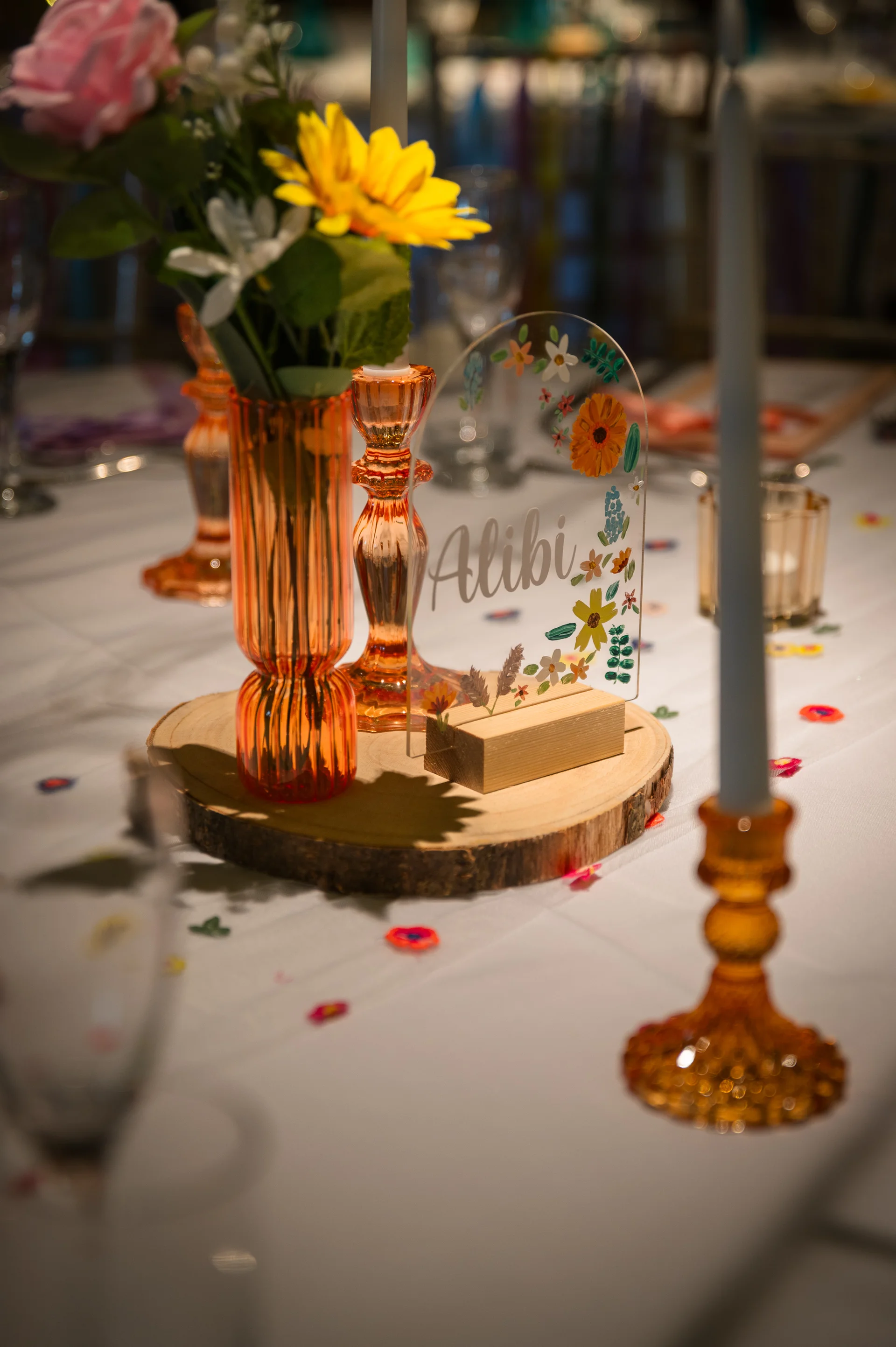 A close up of a colourful wedding tablescape featuring amber glassware, tall taper candles, and a personalised acrylic sign resting on a wooden slice centrepiece.