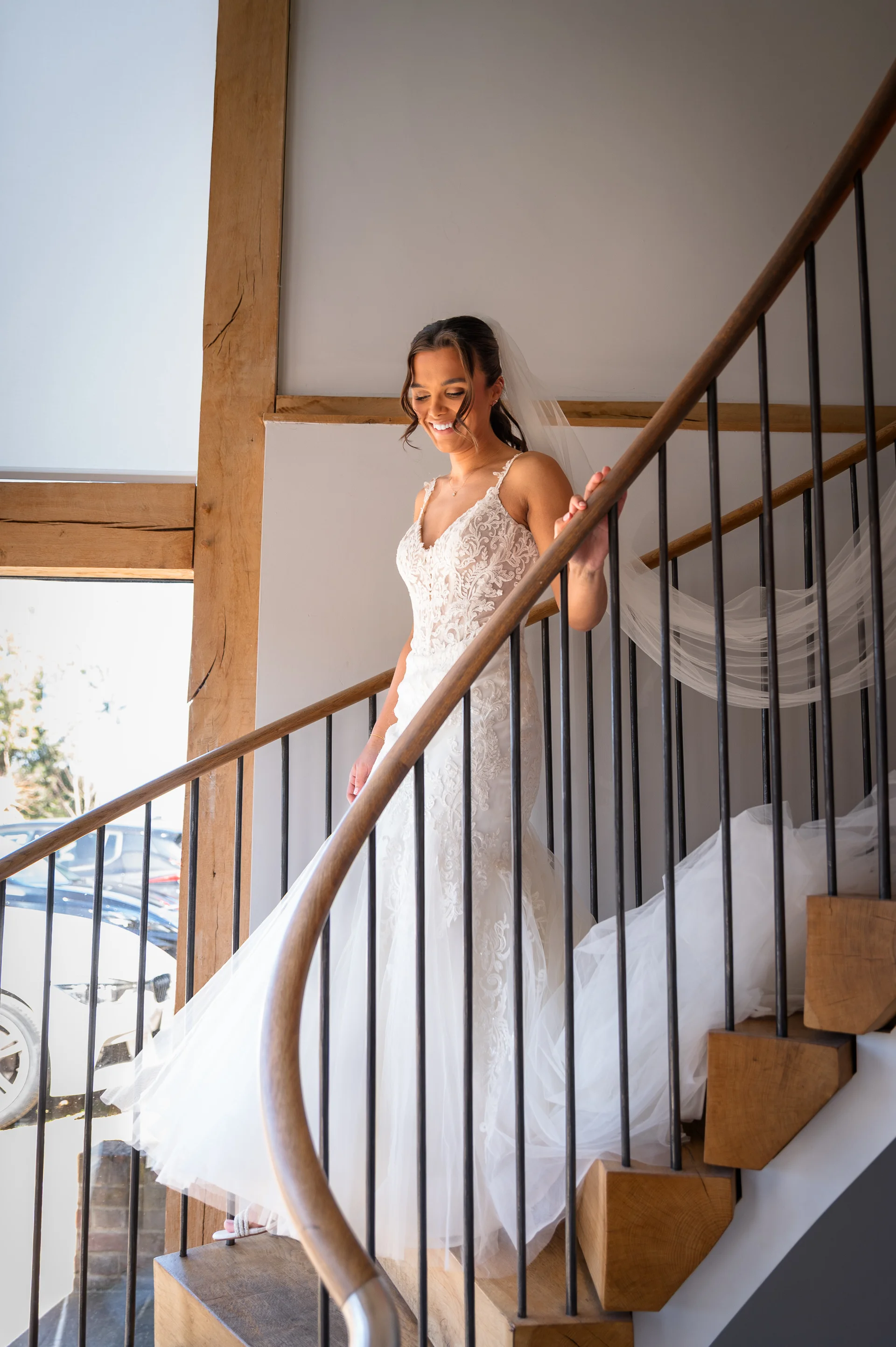 A smiling bride wearing a lace wedding dress with a tulle skirt descends a wooden spiral staircase inside an oak-framed barn.