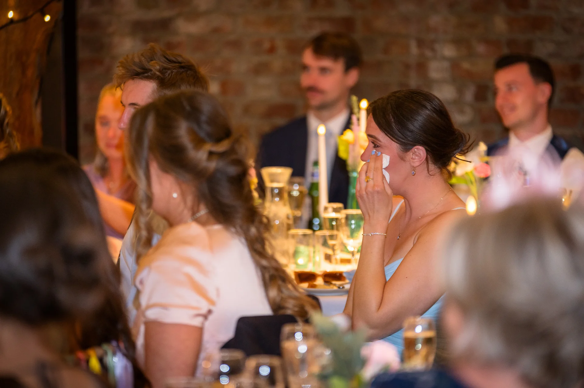 An emotional wedding guest wiping away tears with a tissue during the reception speeches at a candlelit table with exposed brick in the background.