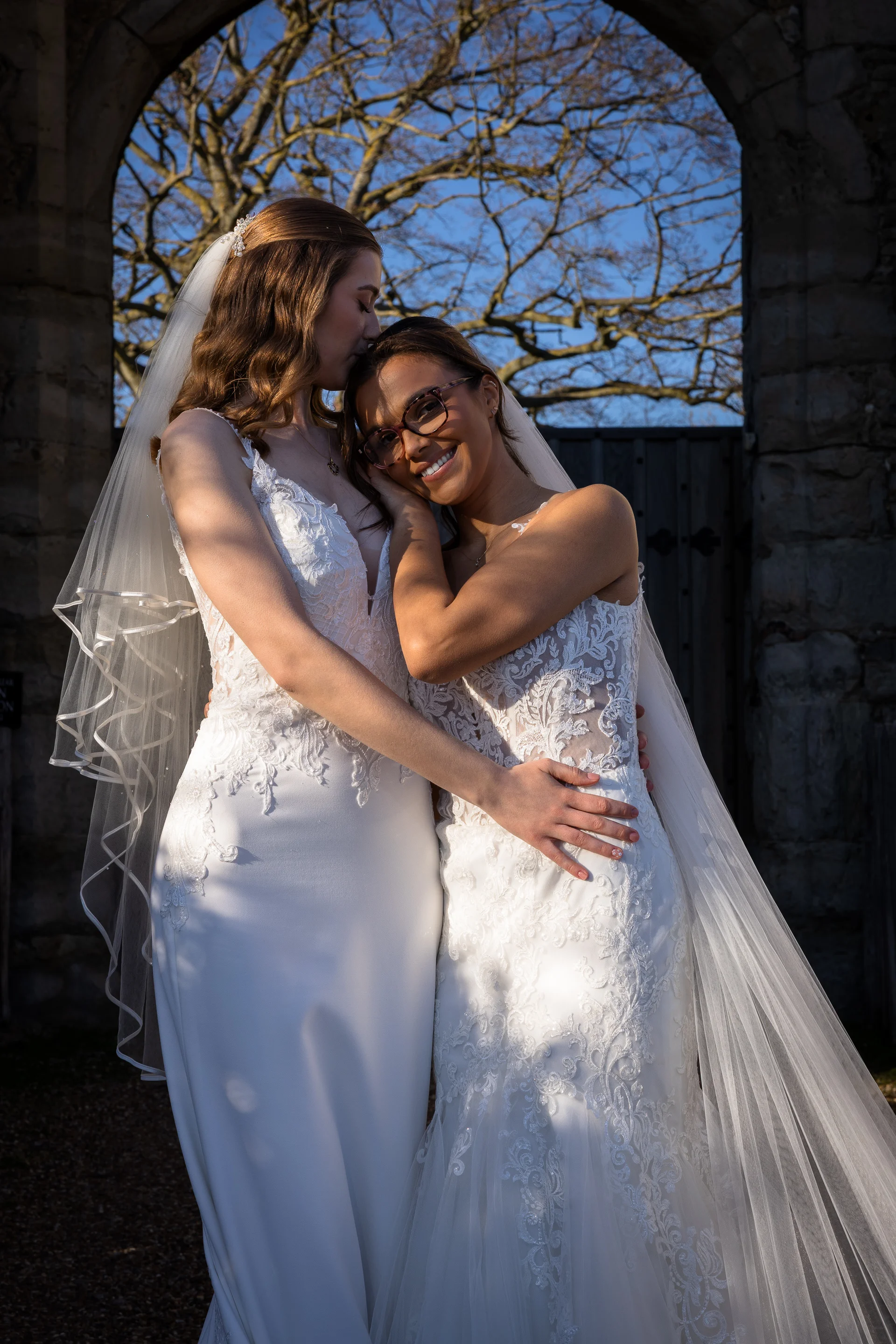 Two smiling brides in lace wedding dresses embrace tenderly under a stone archway during the golden hour.