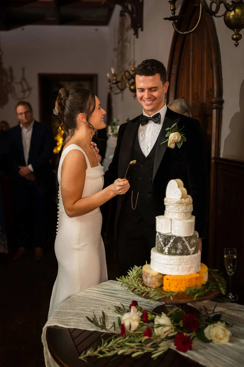 A smiling bride and groom cut into a stacked cheese tower at their evening reception.