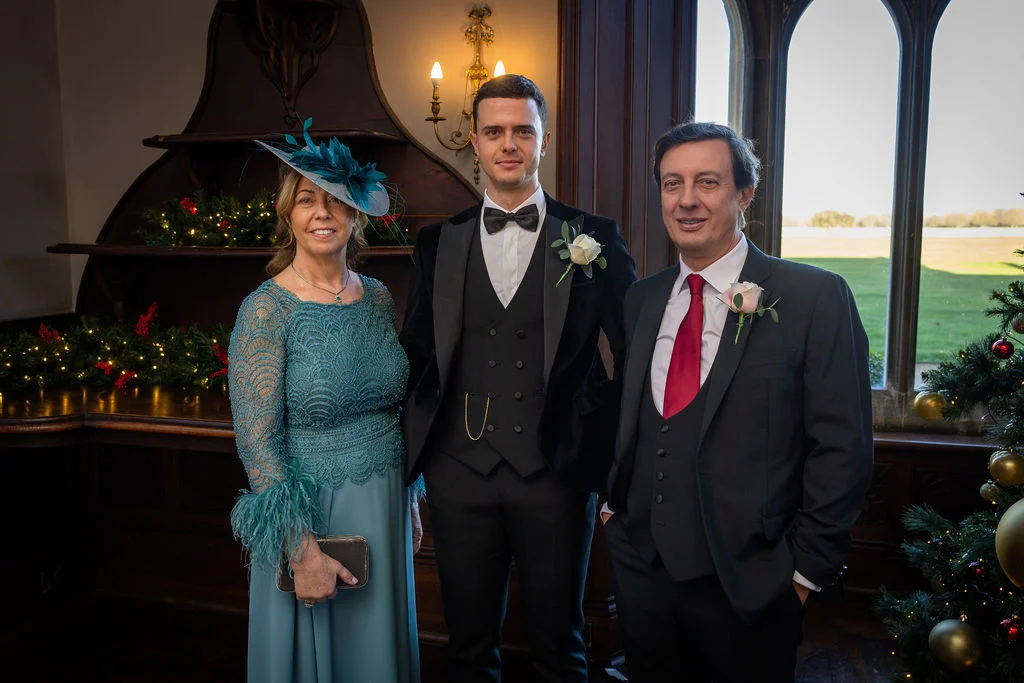 A formal portrait of a groom flanked by his parents inside a country house decorated for a winter wedding.