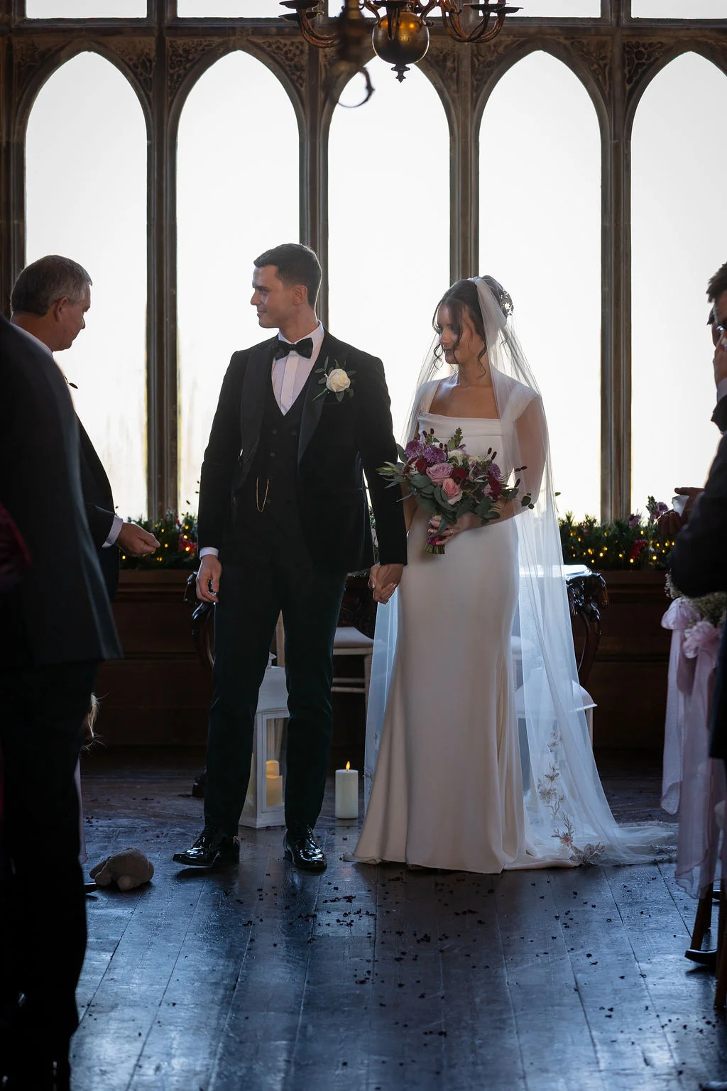 A newly married couple stand hand in hand at the altar in a grand historic hall with tall arched windows, with the groom wearing a black velvet tuxedo and the bride in a sleek crepe gown and embroidered veil.