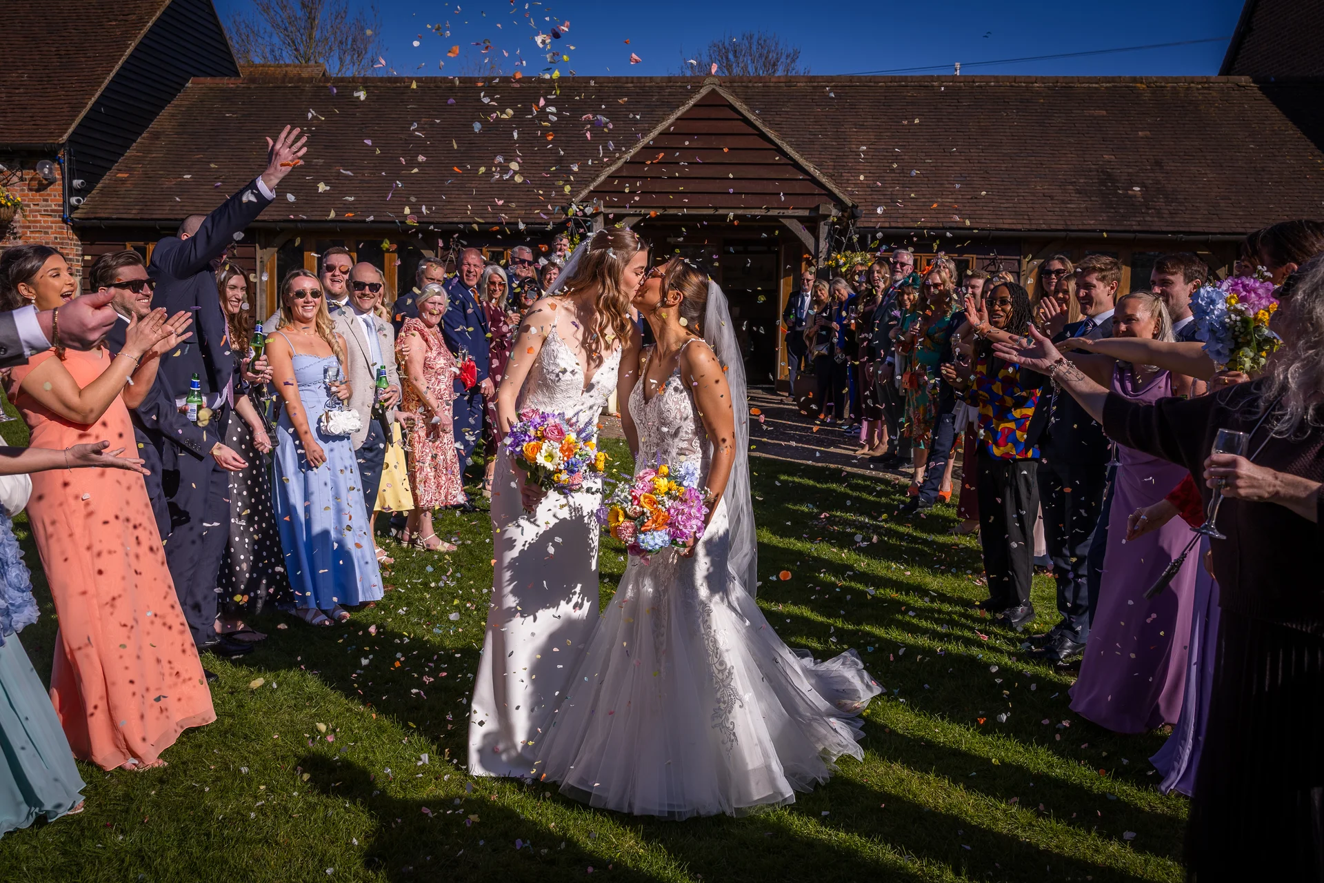 Two brides share a tender kiss as cheering wedding guests shower them with a vibrant flurry of confetti outside a rustic brick barn on a sunny afternoon.