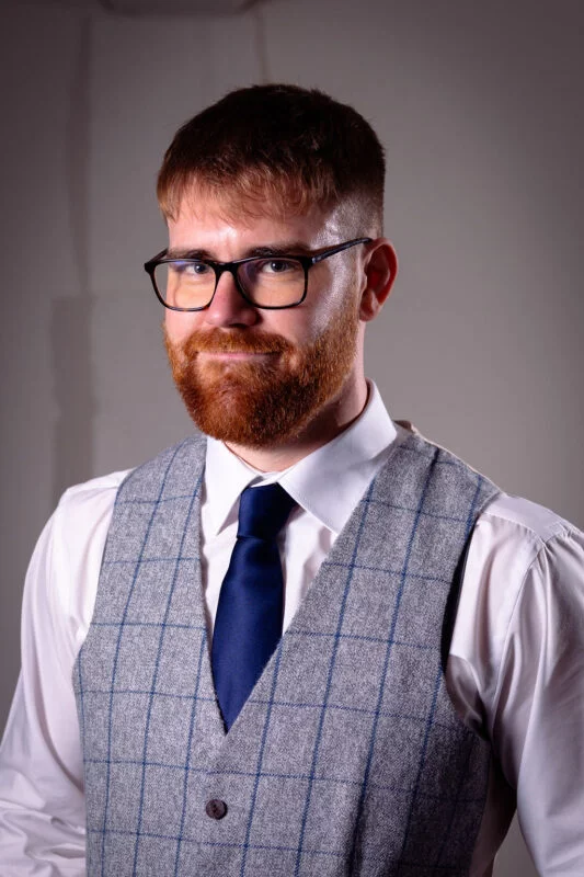 A groom poses for a portrait wearing a grey checked tweed waistcoat, a crisp white shirt, and a blue tie.