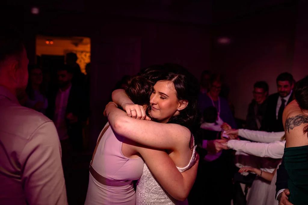 Two brides share a tender embrace on the dance floor during their evening wedding reception bathed in moody coloured lighting.