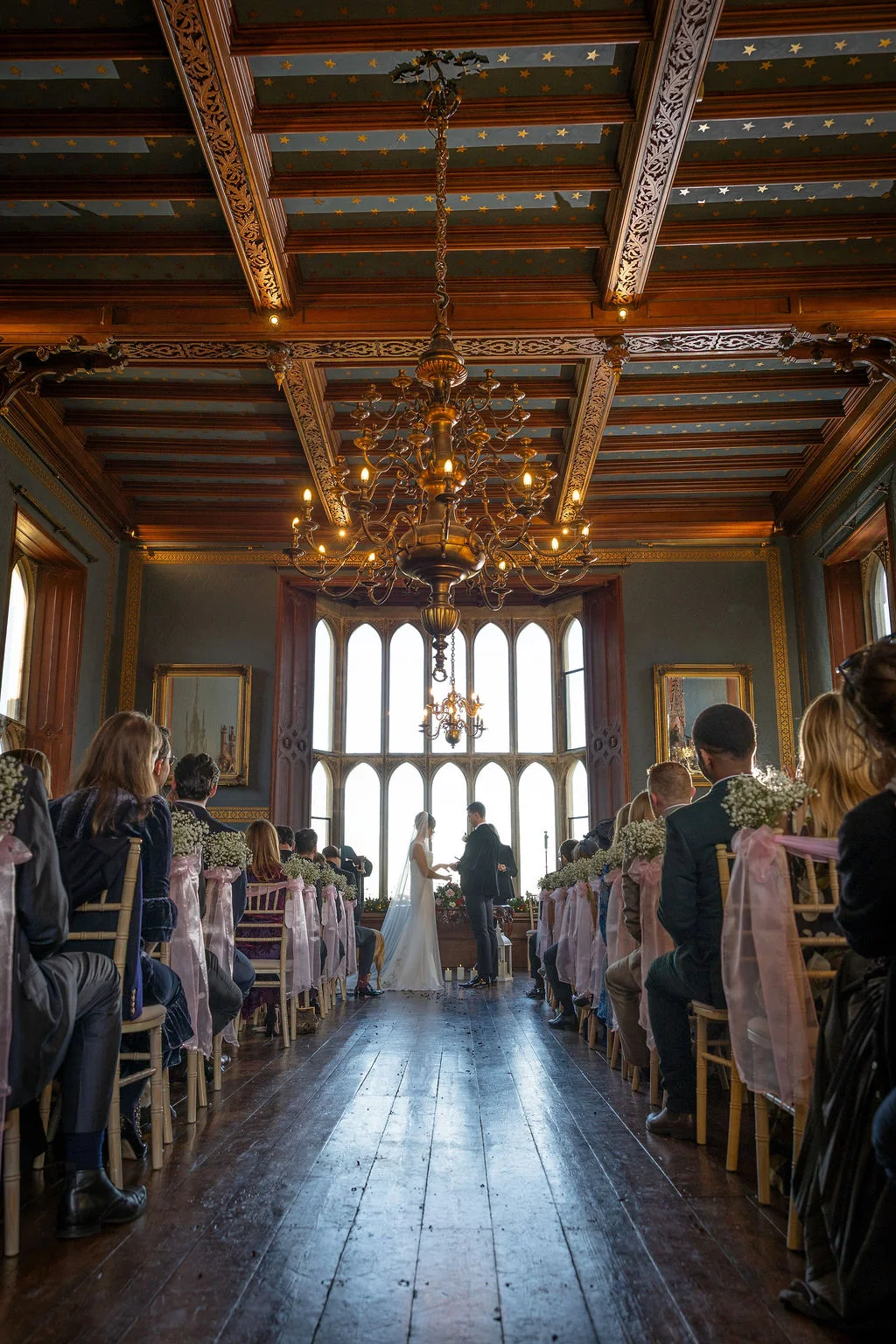 A wide angle shot of a couple exchanging rings during their indoor wedding ceremony in an opulent hall with a vaulted wooden ceiling, striking stained glass windows, and seated guests lining the aisle.