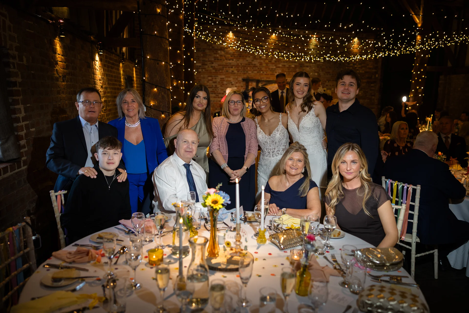 A large wedding party and family members smile together around a candlelit round banquet table inside a rustic barn illuminated by a canopy of fairy lights.