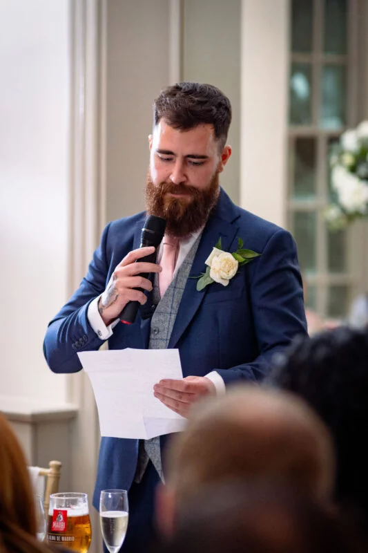 A groom with a full beard delivers his wedding speech, reading notes from a paper while holding a microphone at the reception.