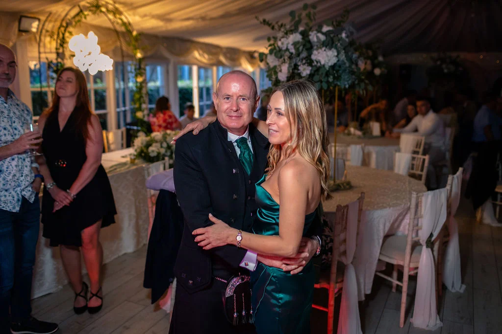 A guest in traditional highland dress wearing a kilt and green tie embraces a woman in a green satin evening gown inside an illuminated marquee wedding reception.