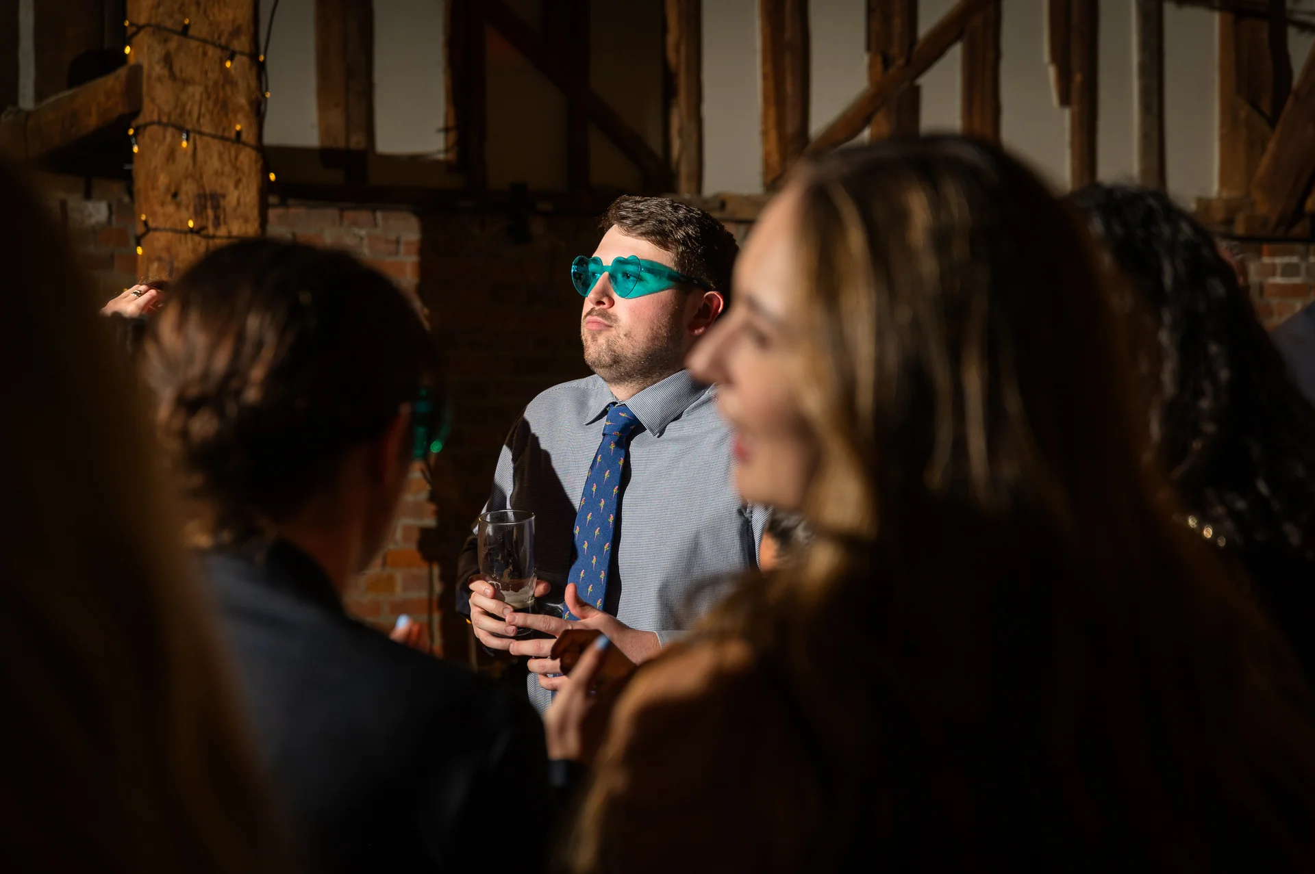 A wedding guest wearing quirky heart-shaped glasses holds a drink during the evening reception inside a rustic barn with exposed brick walls.