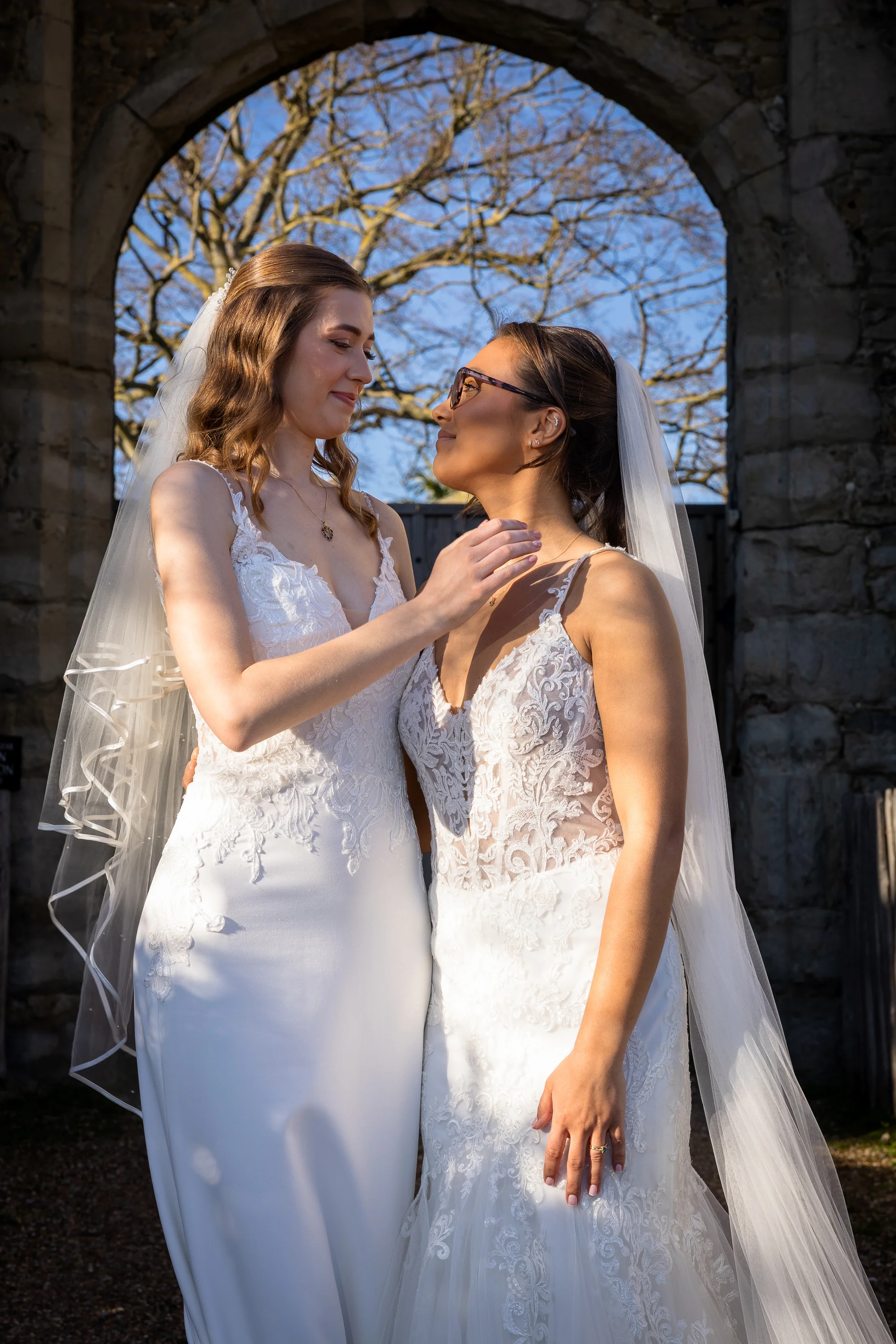 Two smiling brides in lace-accented wedding gowns gaze lovingly at each other while standing framed within an ancient stone archway.