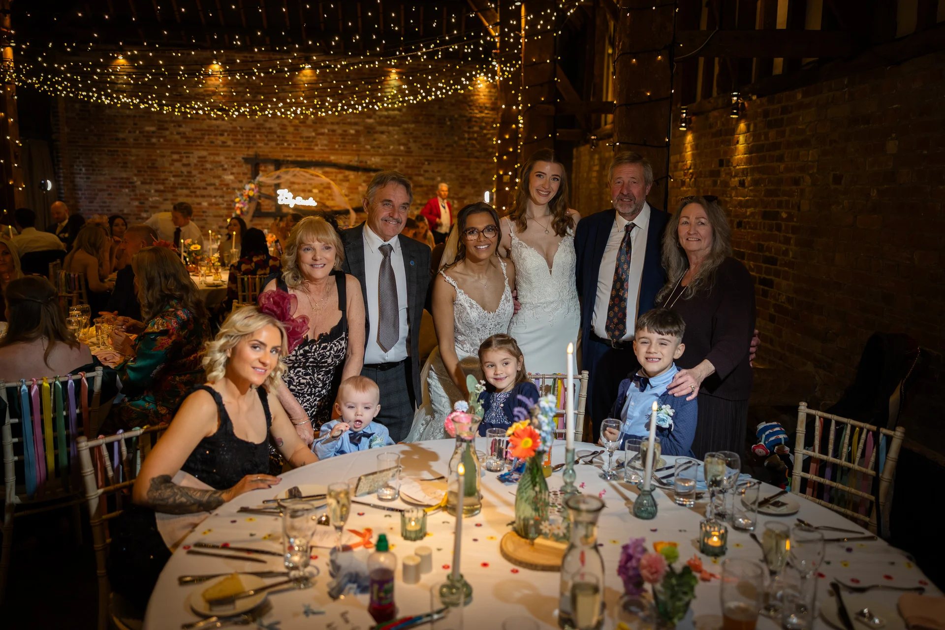 A multi-generational family and wedding party pose together around a candlelit round dining table inside a rustic barn venue illuminated by a fairy light canopy.