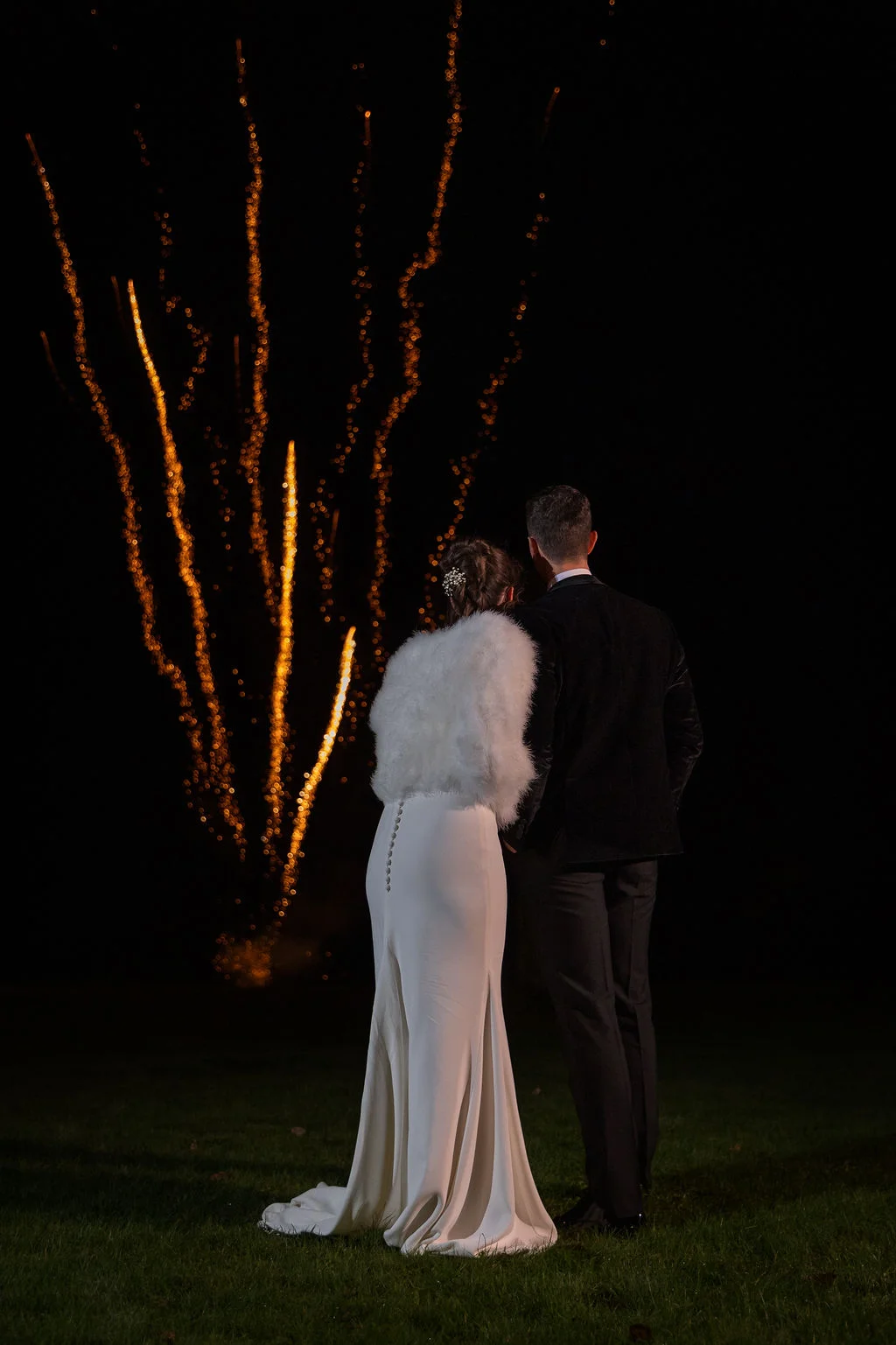A newlywed couple stands outdoors at night, watching a vibrant golden fireworks display light up the dark sky.