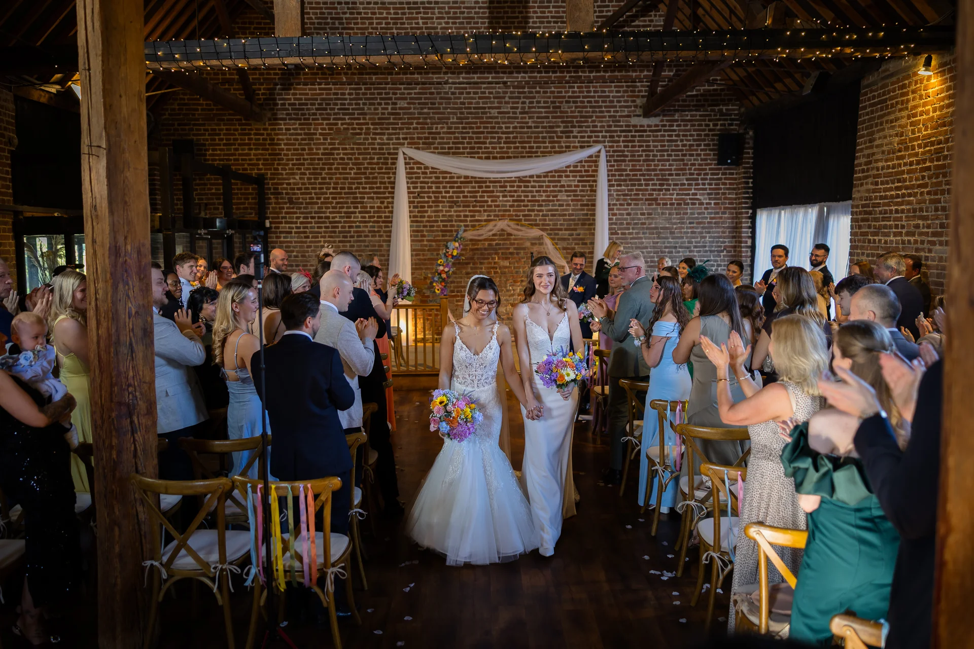 Two smiling brides holding hands and bouquets walk down the aisle together as wedding guests applaud in a rustic brick barn venue.