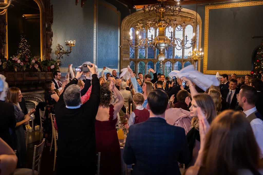 Wedding guests enthusiastically wave their napkins in the air while cheering during speeches inside a grand, wood-panelled stately home ballroom.