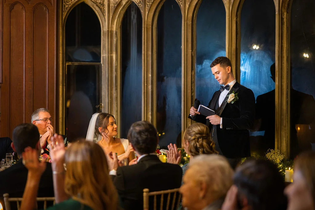 A groom in a black velvet tuxedo delivers his speech while holding notes and a microphone, as the bride and seated guests look on attentively inside an elegant venue with large arched windows.