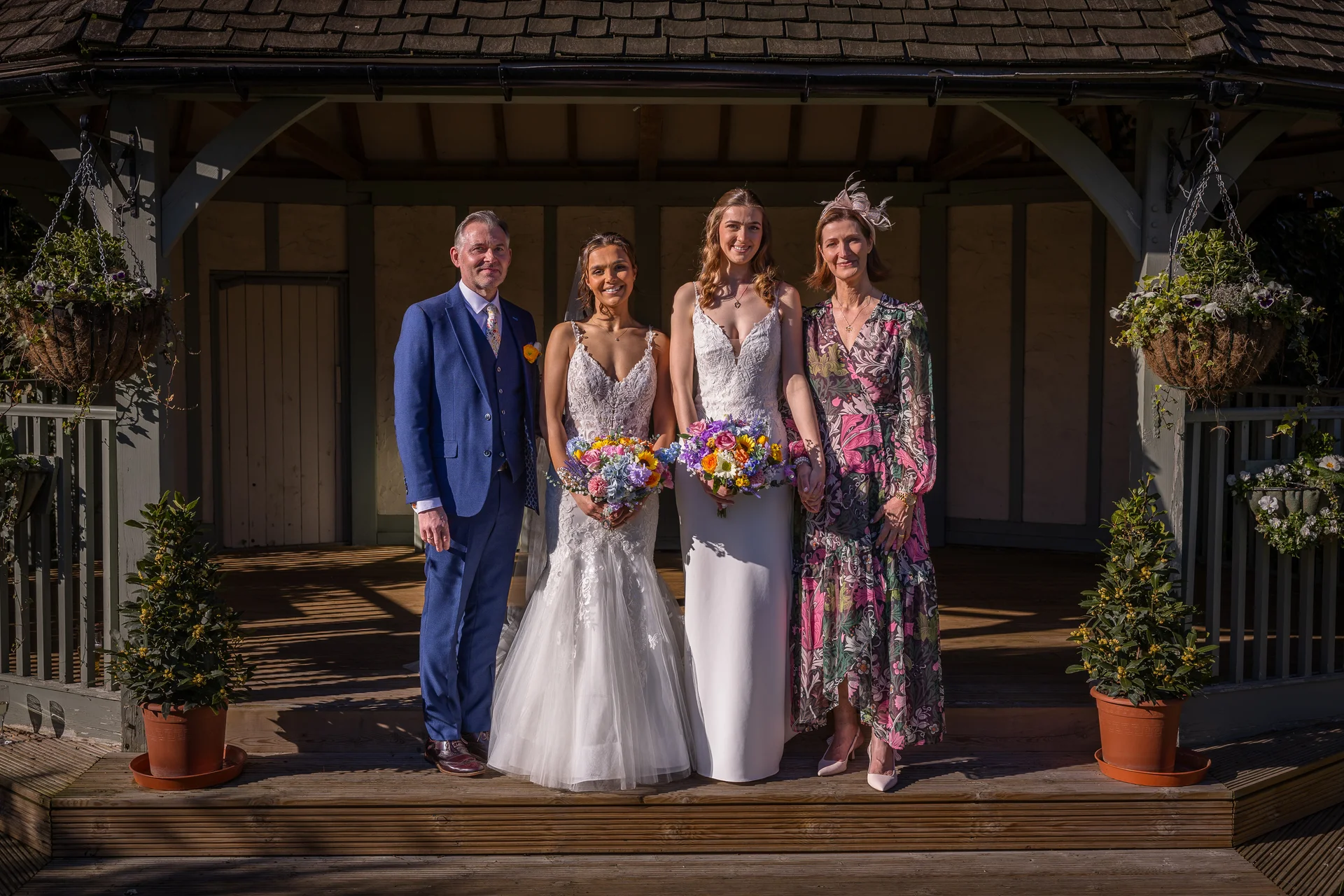 A formal group portrait of two brides, a man in a blue suit, and a woman in a floral dress standing on the wooden steps of a bandstand outdoors in bright sunlight.
