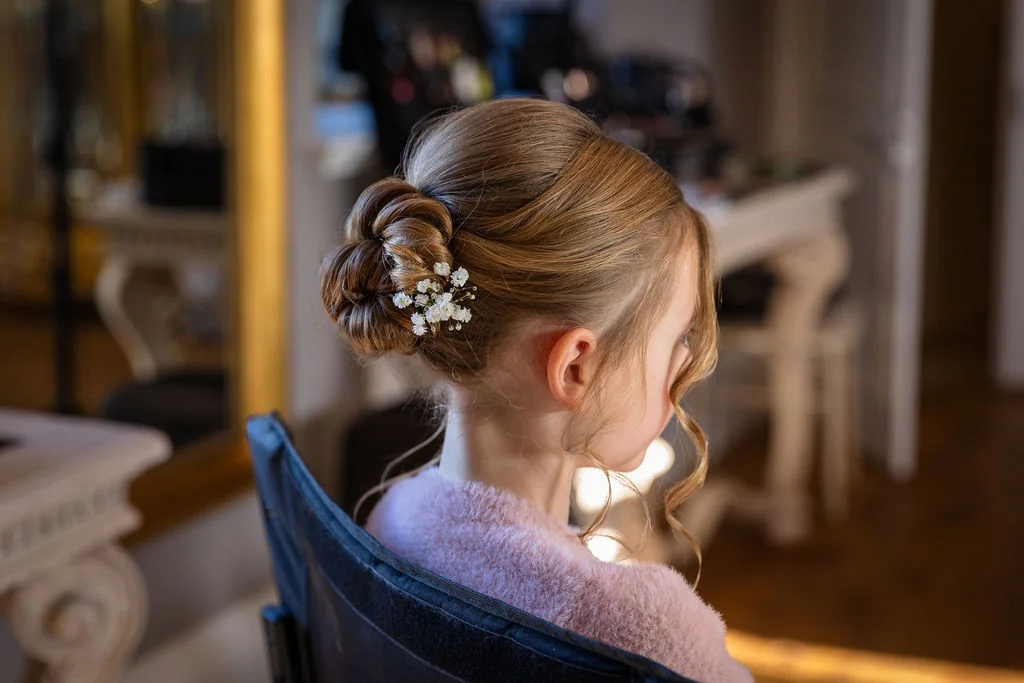 A close-up back view of a bride sitting in a makeup chair showcasing an intricate updo adorned with delicate baby's breath flowers and soft face-framing tendrils.