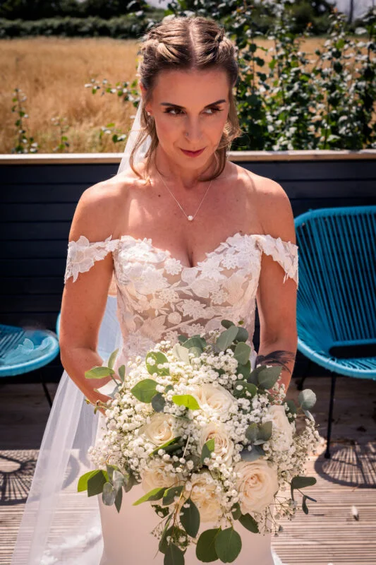 A stunning bride holds a lush white rose and baby's breath bouquet, wearing an off-the-shoulder lace gown with a sweetheart neckline in the bright sunshine.