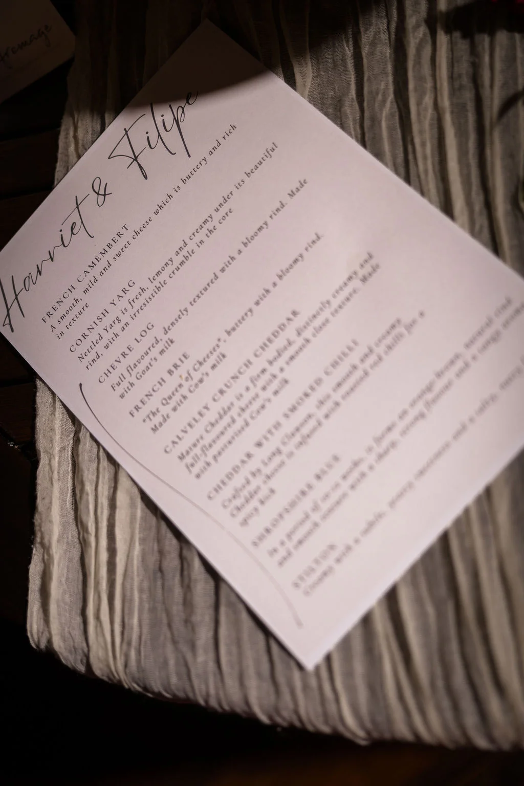 An elegant wedding cheese menu card featuring calligraphy names sits atop a textured neutral linen table runner.