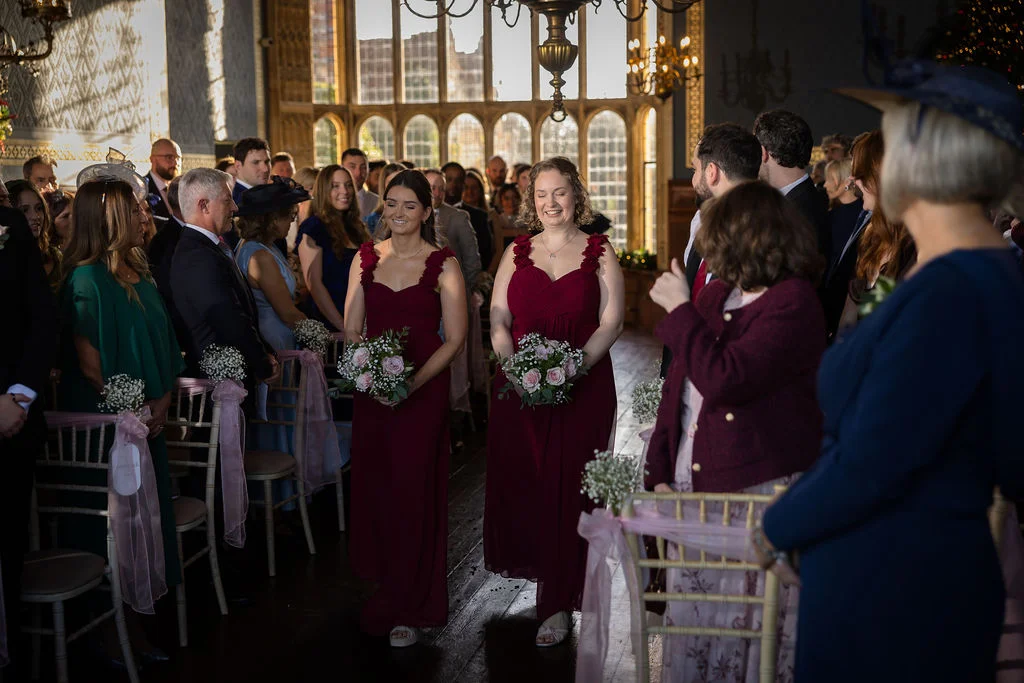 Bridesmaids in burgundy dresses walking down the aisle holding floral bouquets during a wedding ceremony inside a historic venue.