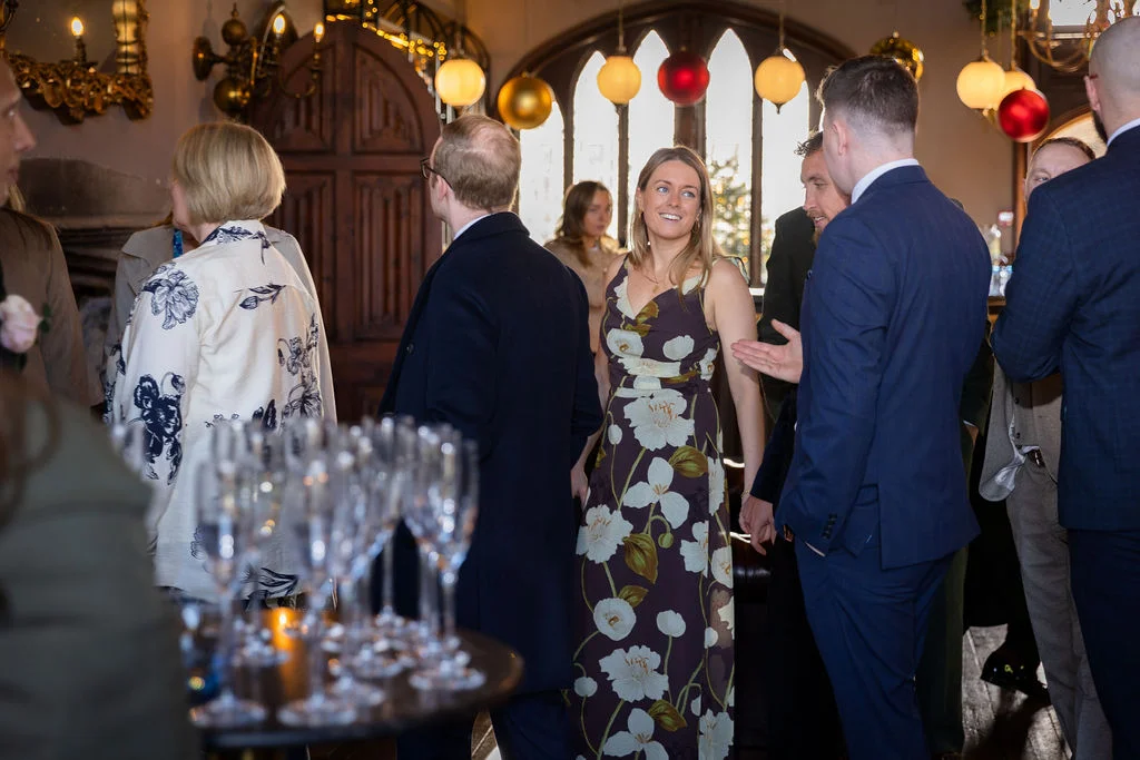 Wedding guests mingle happily during the drinks reception inside a historic venue decorated with festive baubles and large windows.