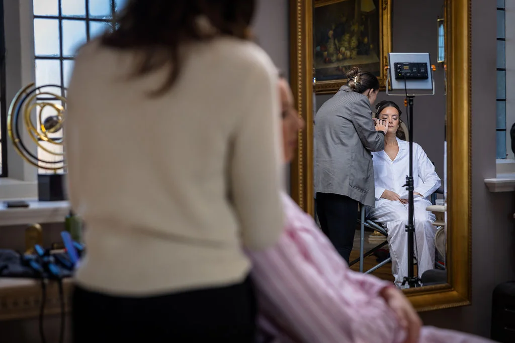 A bride having her makeup applied during morning bridal preparations is captured through a large ornate mirror reflection.