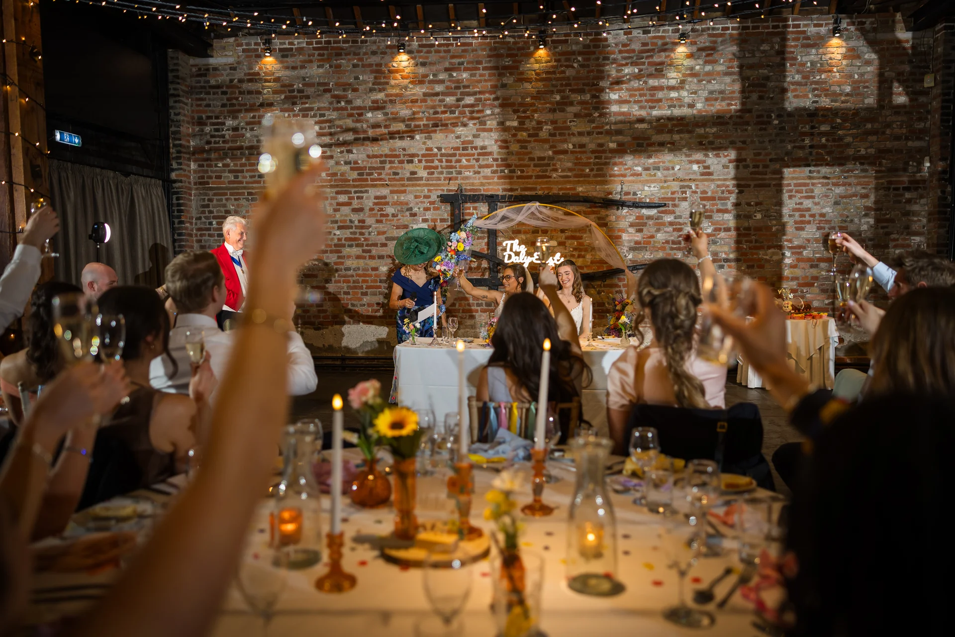 Wedding guests raise their glasses in a toast during the speeches at a warehouse reception featuring exposed brick walls and twinkling fairy lights.