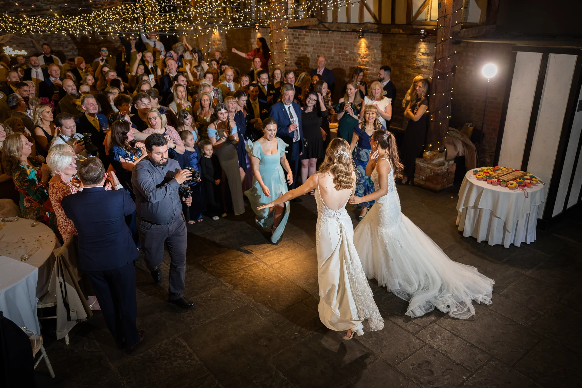 Two brides share their first dance in a rustic stone barn adorned with a twinkling fairy light canopy while guests look on enthusiastically.