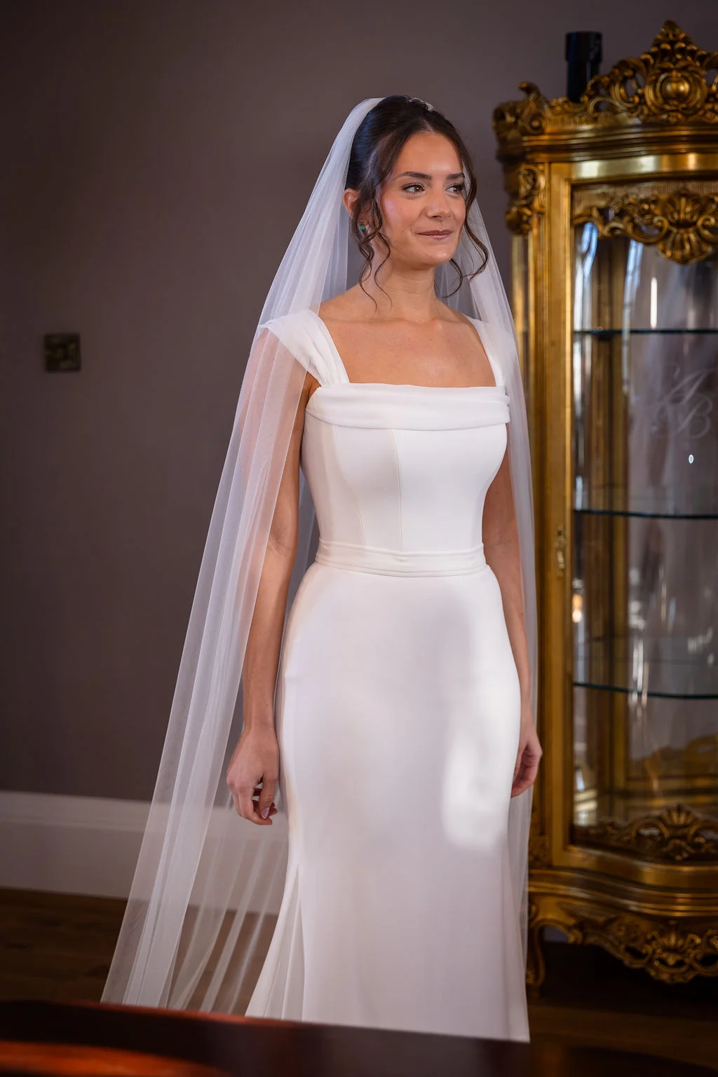 A graceful solo bride wearing a clean square-neckline crepe gown and a long veil stands proudly indoors beside an ornate golden cabinet.