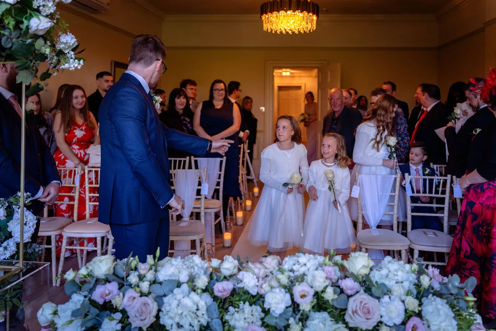A groom in a navy suit directs two young flower girls holding white roses down the aisle during an indoor wedding ceremony.