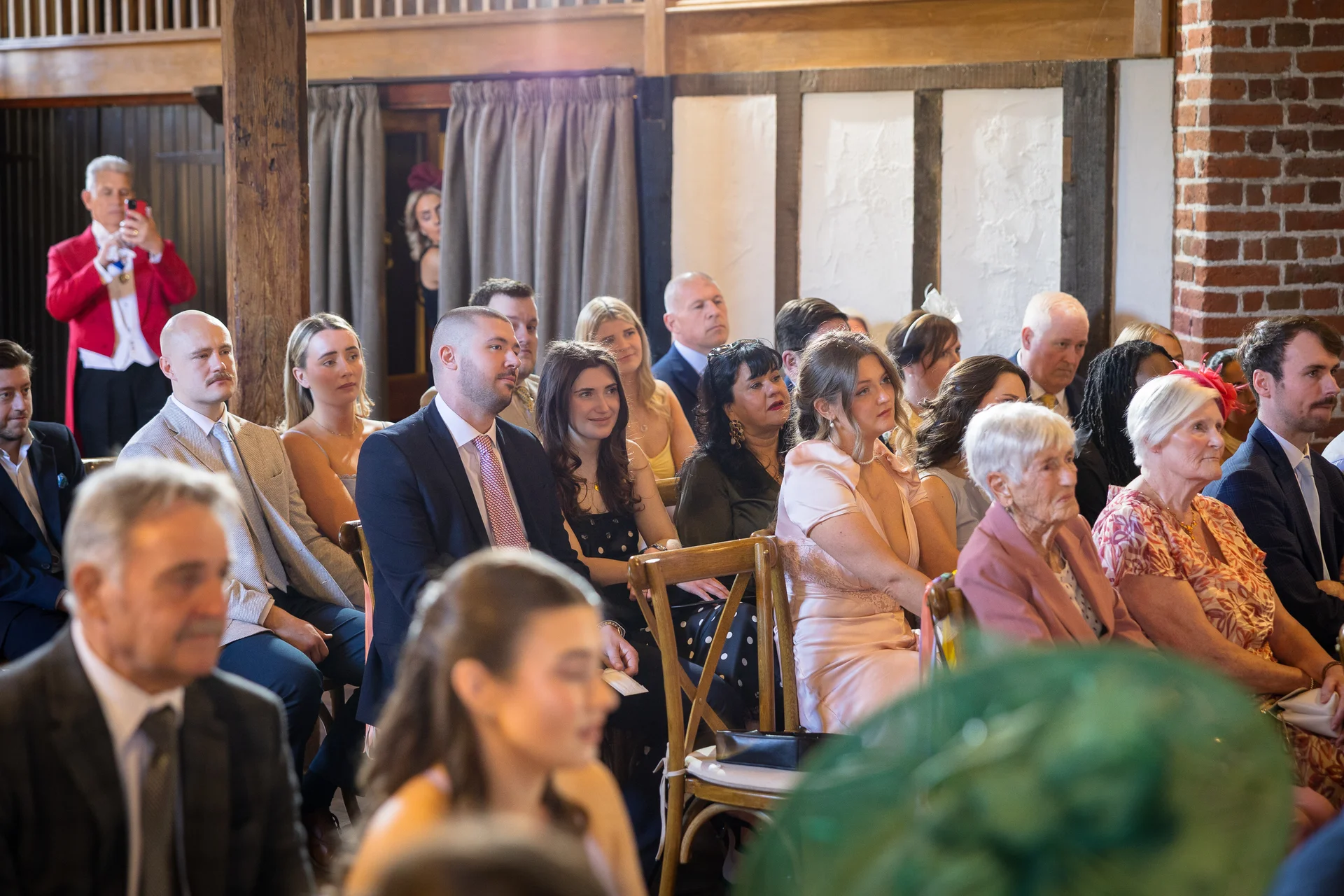A candid wide shot captures a diverse gathering of wedding guests seated inside a rustic barn venue with exposed brick and wooden beams.
