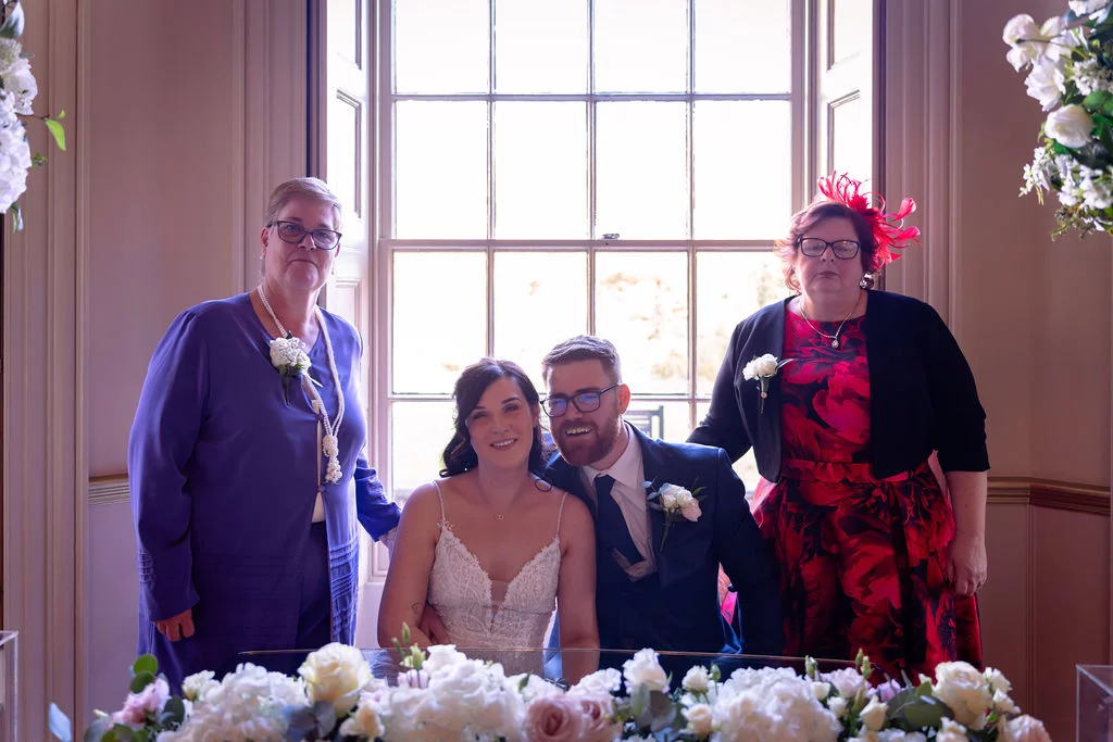 The bride and groom pose smilingly alongside two family members behind a flower-adorned table in front of a large bright window.