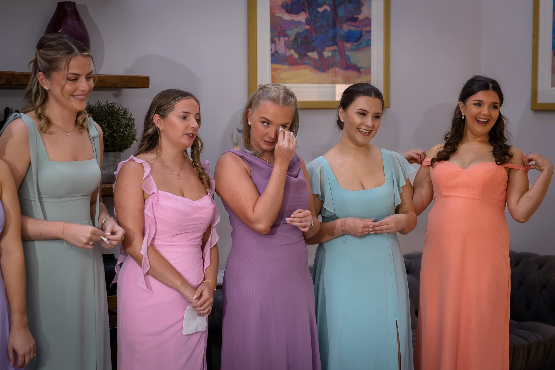A group of bridesmaids in colourful pastel dresses react emotionally during wedding preparations, with one wiping a tear.