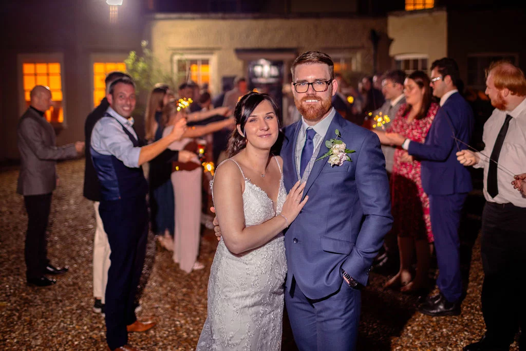 A bride in an embellished gown and groom in a navy suit pose together at night outdoors, framed by wedding guests holding lit sparklers.