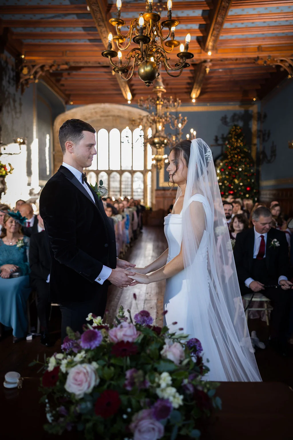 A bride and groom hold hands during their indoor ceremony inside a grand stately home featuring ornate chandeliers and tall windows.