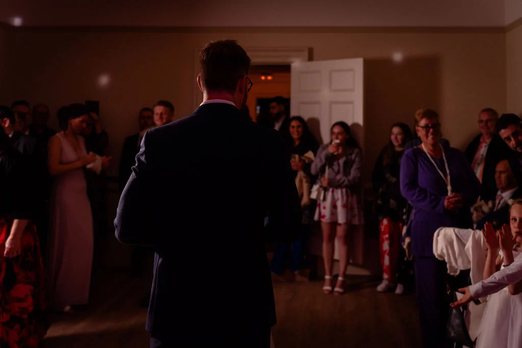 A groom stands with his back to the camera delivering a speech to an attentive audience of wedding guests gathered indoors.