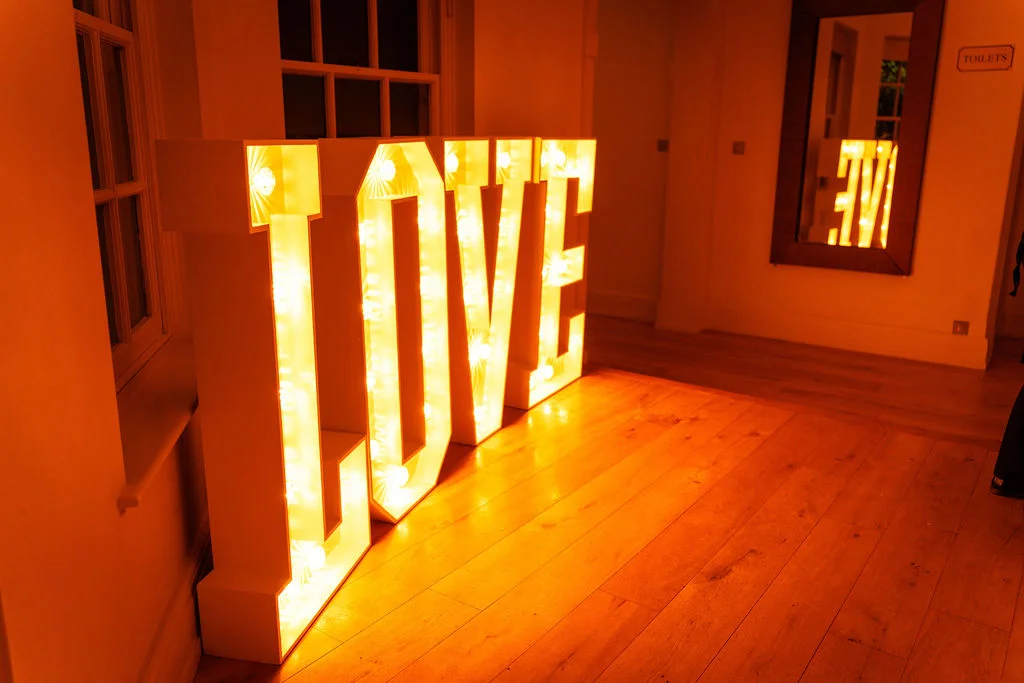Large illuminated love letters cast a warm, glowing amber light across the wooden floor inside a venue hallway, reflected in a nearby wall mirror.