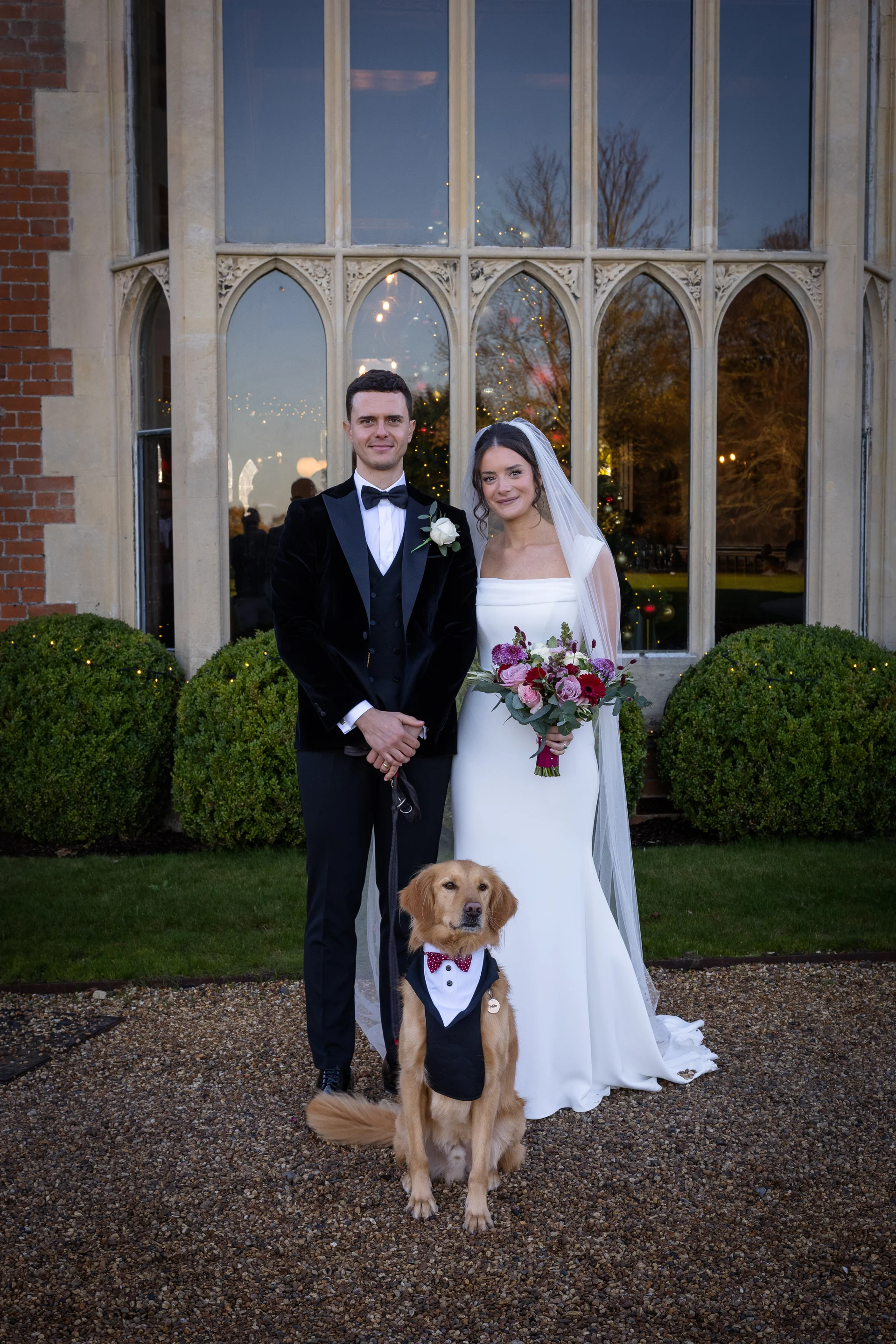 A newlywed couple poses outside a grand manor house with their golden retriever wearing a tuxedo and bow tie.