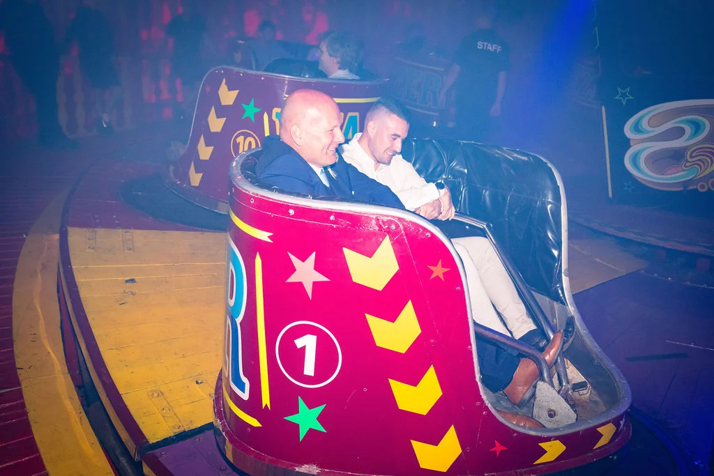 Two male guests enjoy a ride on a brightly decorated red and yellow dodgem car at a funfair-themed wedding evening reception.