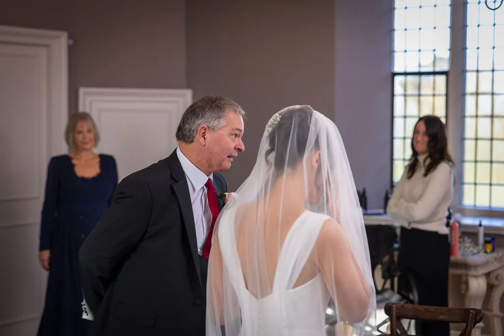 A father shares an emotional first look with his bride daughter wearing a white V-neck gown and veil, while family members look on indoors.