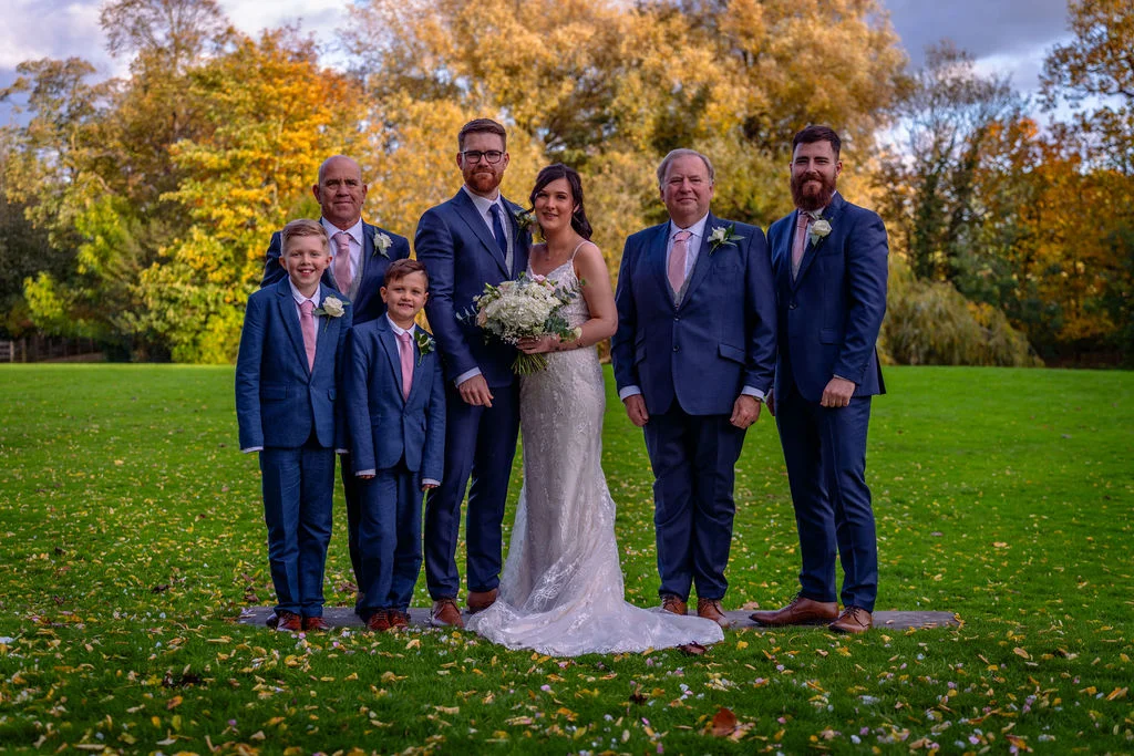 A bridal party posed outdoors on a lush green lawn with autumn trees in the background during an autumn wedding.
