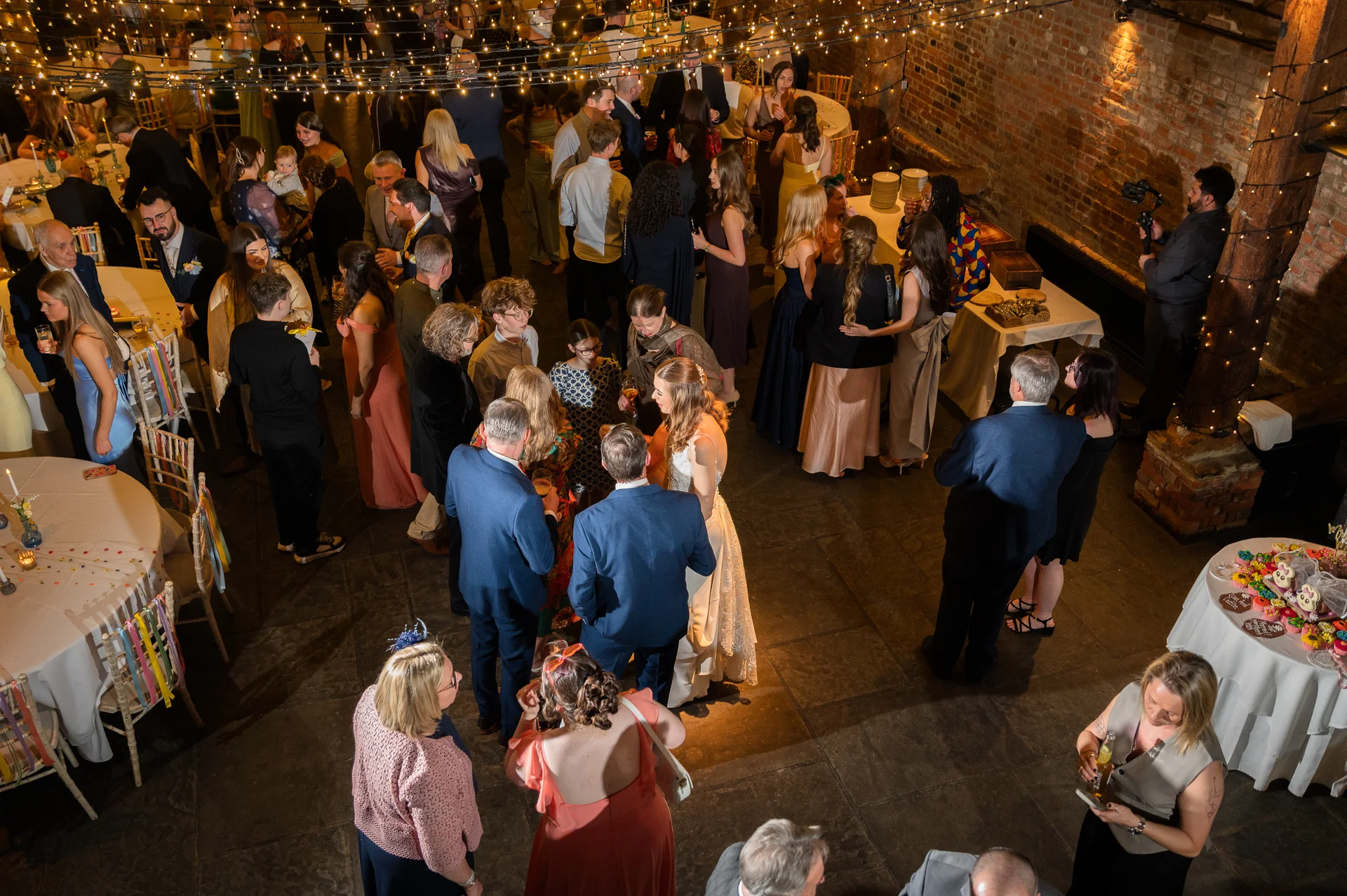 An elevated view of a lively evening wedding reception inside an industrial brick venue illuminated by a magical canopy of fairy lights.
