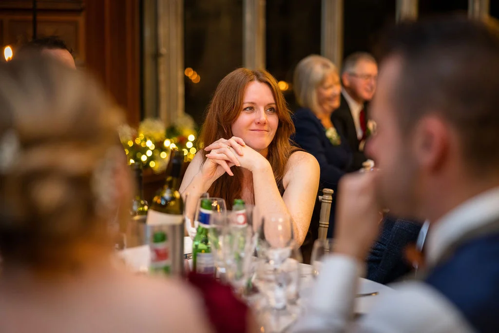 A female wedding guest with auburn hair smiles warmly as she leans on her folded hands at a banquet table during the reception.
