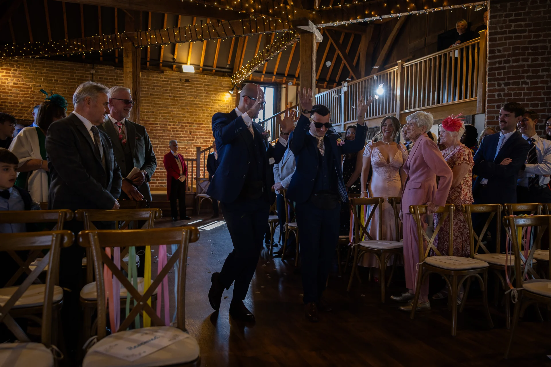 Two grooms in matching navy suits raise their arms joyfully as they walk down the aisle amidst cheering guests inside a rustic barn venue.