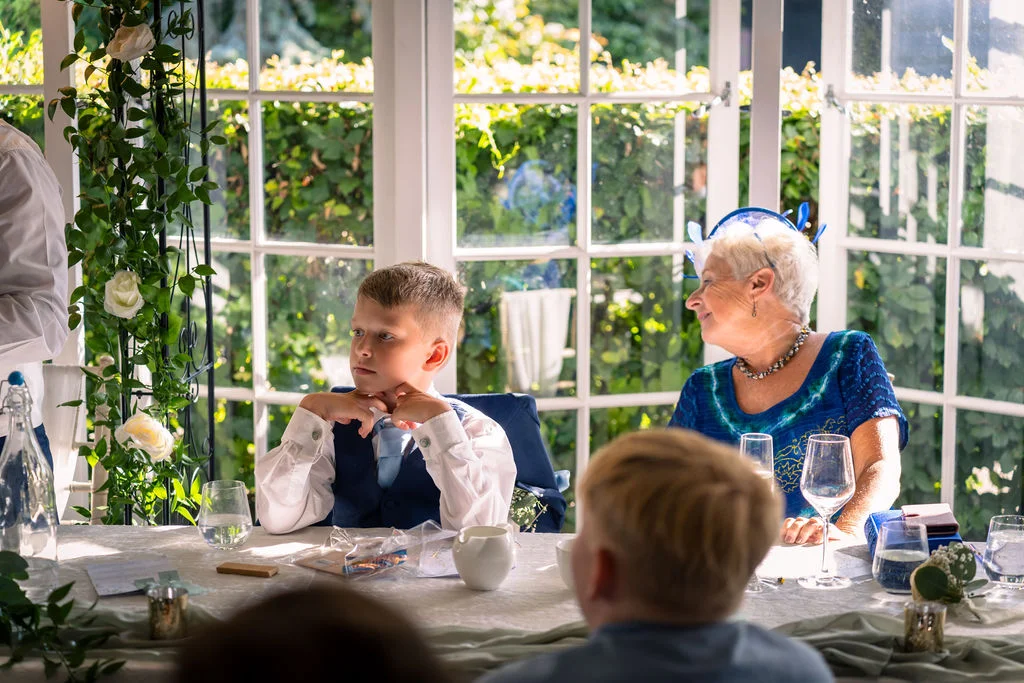 A young page boy in a waistcoat sits at a wedding breakfast table next to a laughing older guest near bright conservatory windows.