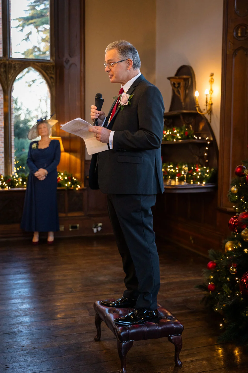 A man in a dark suit delivers a wedding speech holding a microphone and notes while standing on a leather footstool in a wood-panelled room decorated for Christmas.