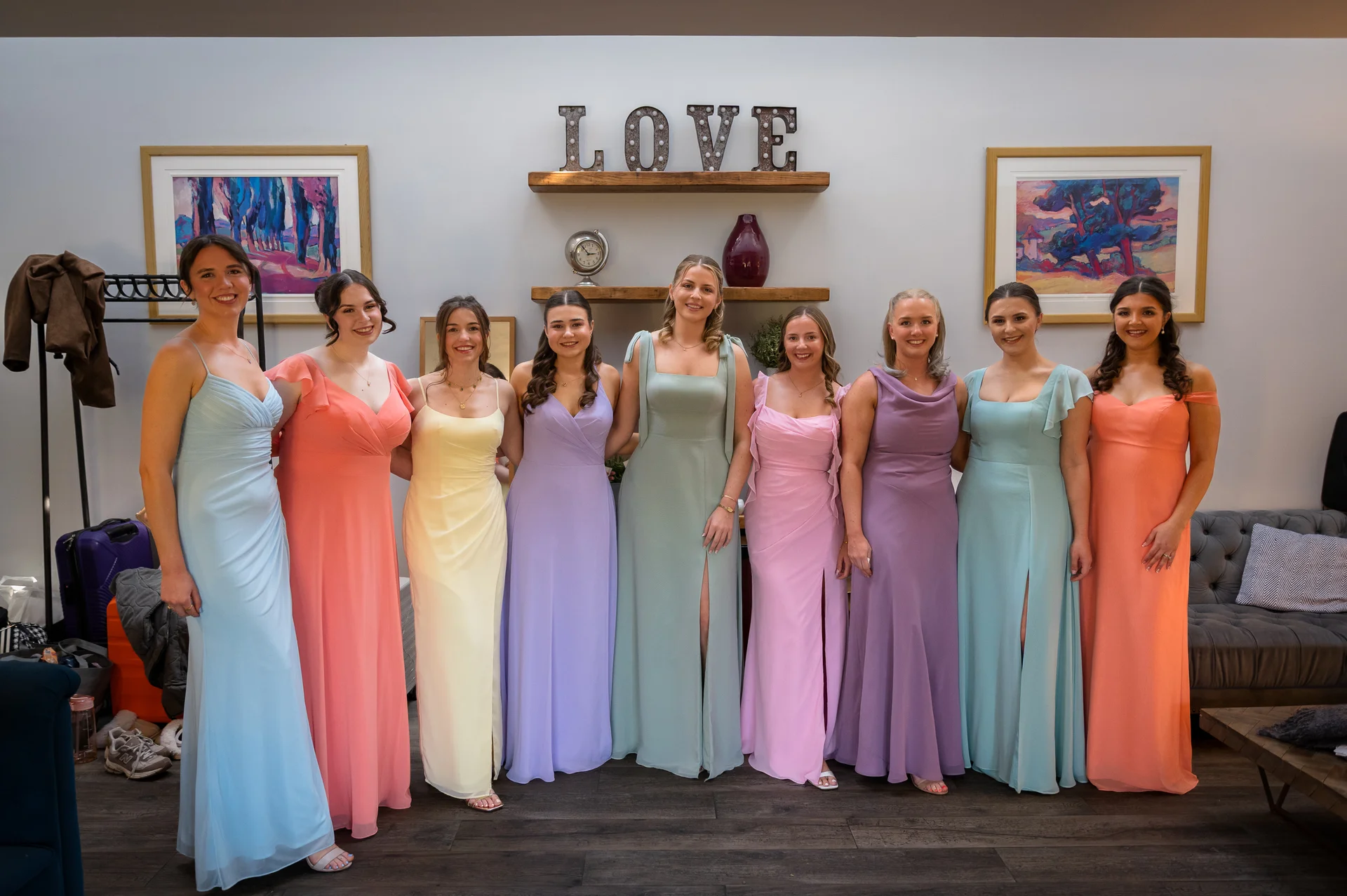 A group of nine smiling bridesmaids pose together wearing mismatched pastel-coloured floor-length dresses in pastel shades of blue, peach, yellow, lilac, green, pink, and purple against a white wall with framed artwork.