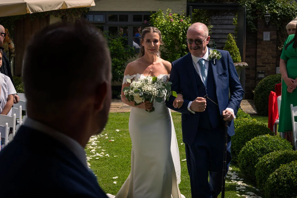 A smiling bride walks down an outdoor garden aisle holding a white floral bouquet, linked arm-in-arm with her father as she approaches the groom.